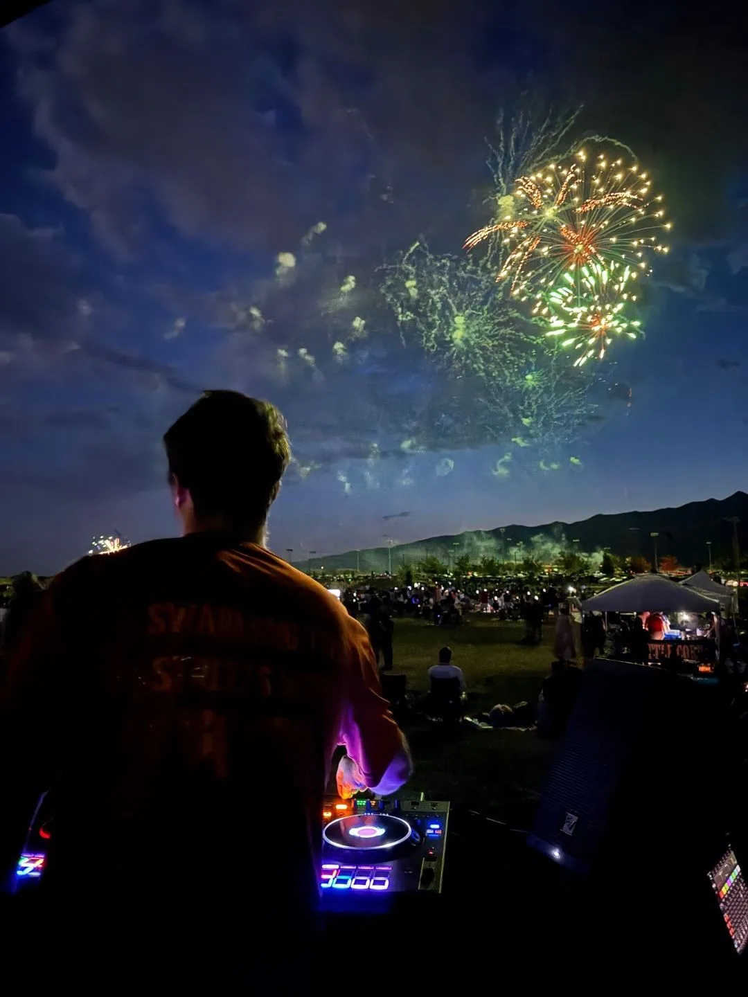Fireworks exploding over a large crowd at night with DJ in foreground