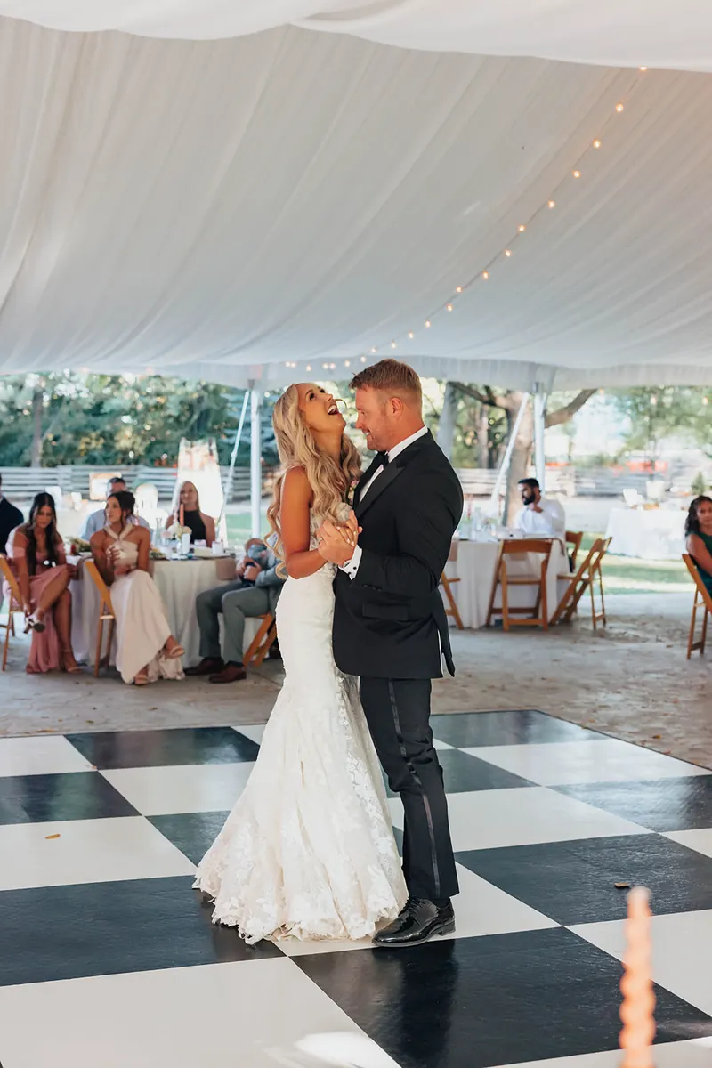 Couple sharing their first dance on a black and white checkerboard floor under a tented reception