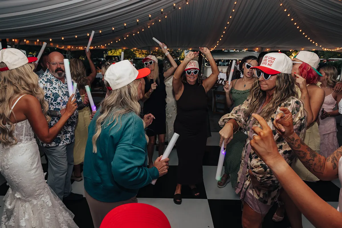 A packed wedding dance floor under tent string lights, guests waving foam light sticks