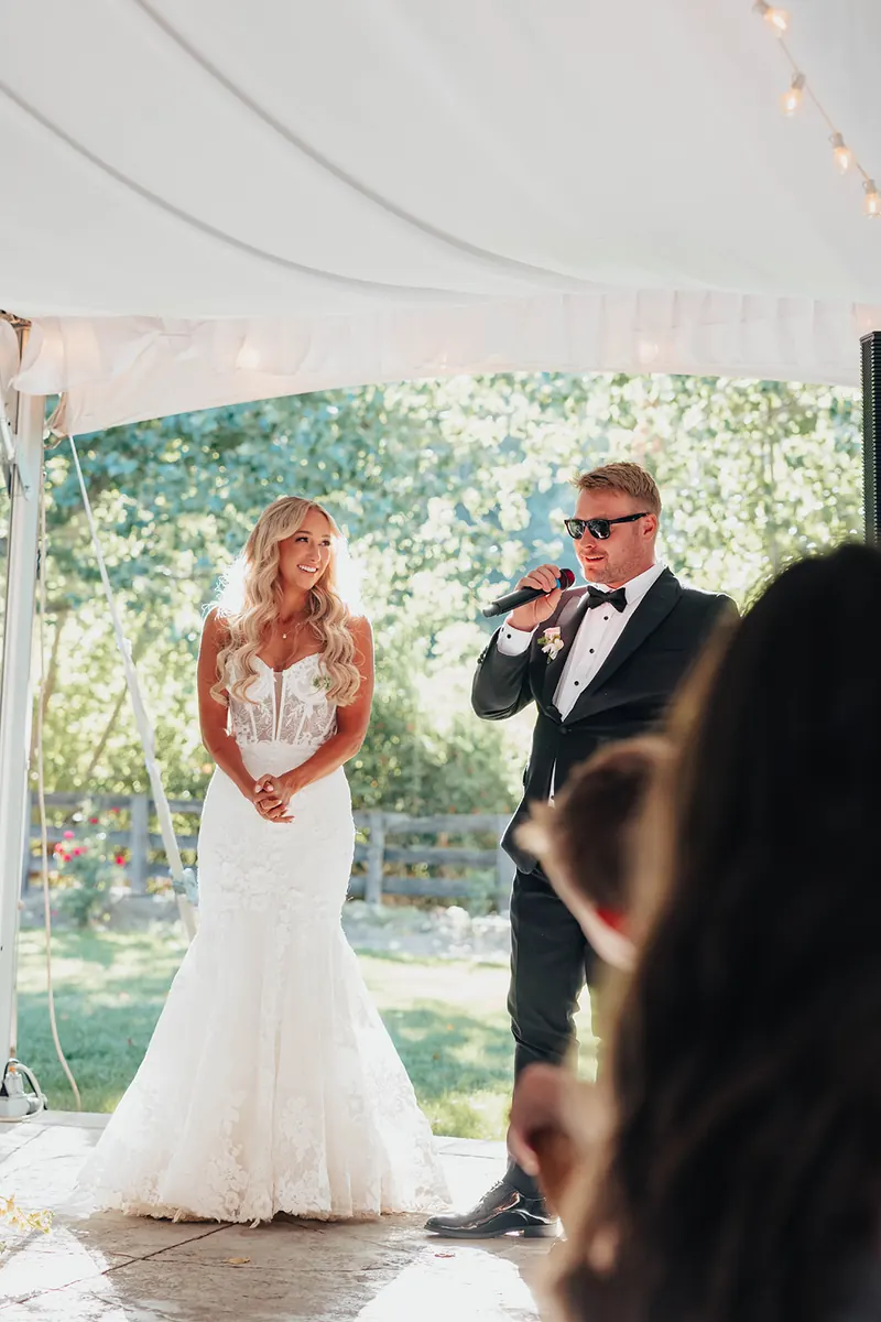 Bride listening as the best man gives his toast on a wireless mic under a white reception tent