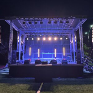 An empty outdoor stage glows with purple and yellow lights, ready for any event with the Concert Level Audio Package - Treasure Valley Rental. Trusted speakers flank the stage, set on a grassy area beneath the night sky—perfect for performances or gatherings.